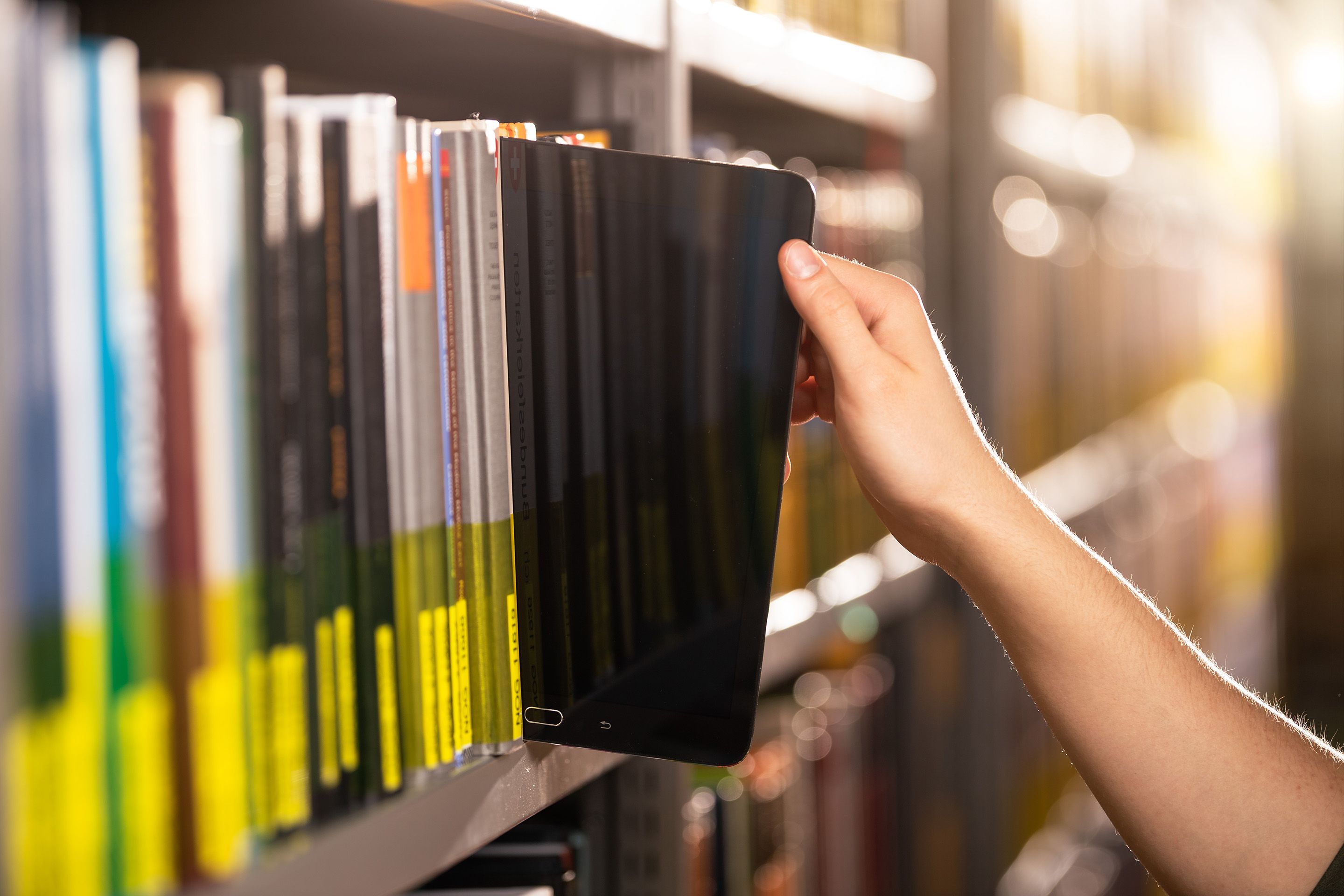 Auf dem Bild sieht man eine Hand, die ein Tablet aus dem Bücherregal zieht.
