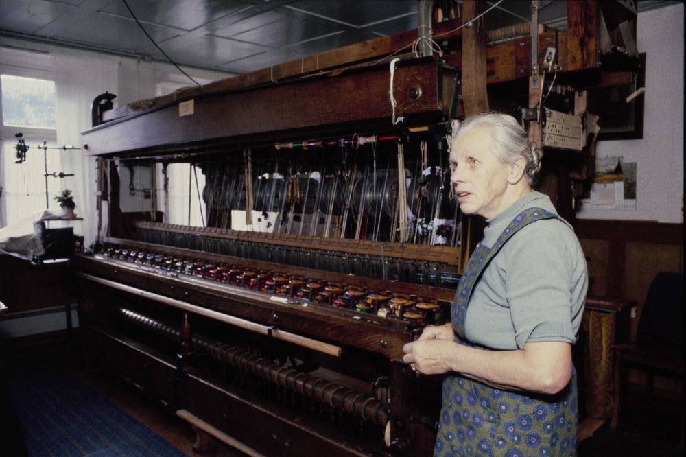 Das Bild zeigt die Heimposamenterin Lisette Waldner neben ihrem Webstuhl, aufgenommen 1981.