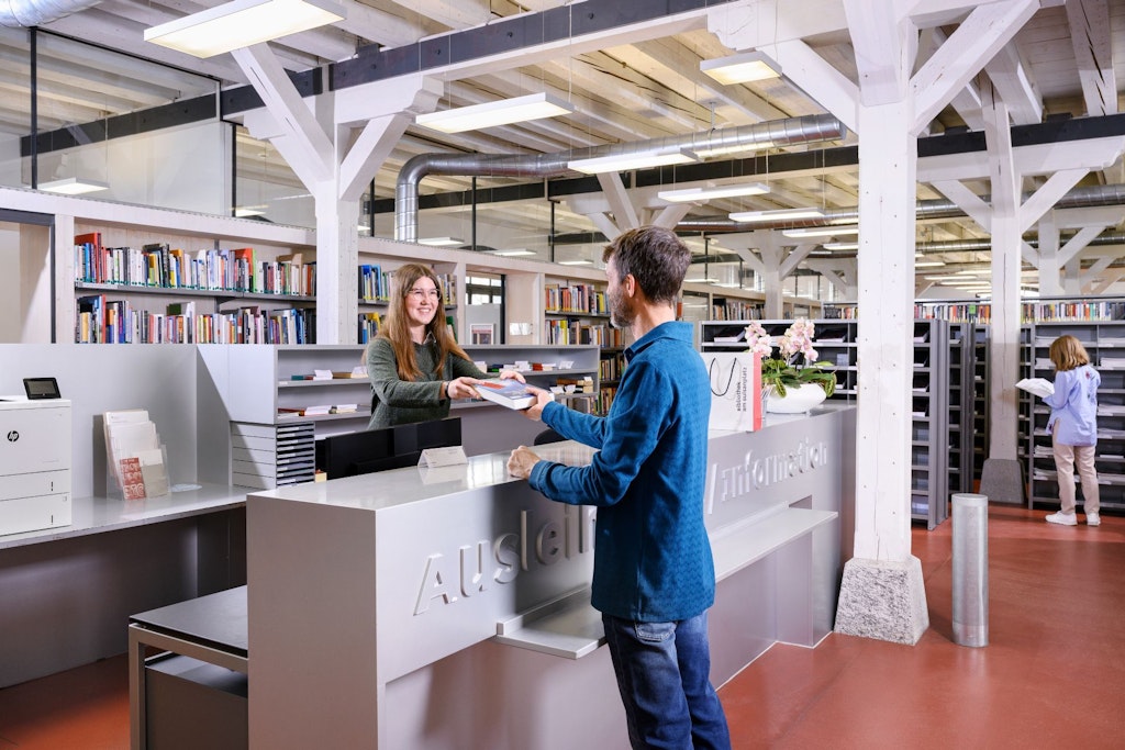 Auf dem Bild sieht man eine Lernende, die einem Bibliotheksmitarbeiter Bücher aushändigt.