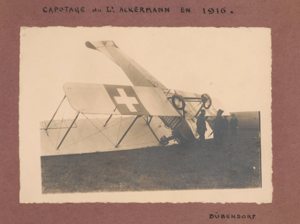 Des hommes devant un avion qui se retrouve sens dessus dessous après avoir piqué du nez. Le capotage de l’avion du lieutenant Ackermann s’est produit en 1916 à Dübendorf, d’après ce qui est noté sur la photographie (éditeur : [probablement] Hans Bandi. Accidents d’aviation, 1916-1918. BIG DU 549).